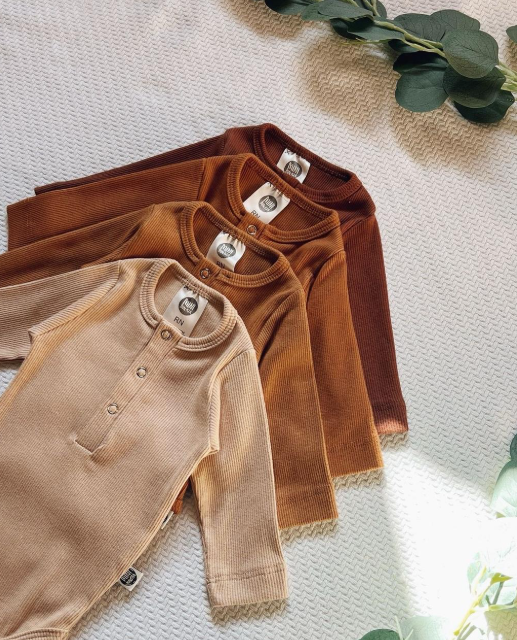 Macacões de bebê em tons terrosos organizados em degradê sobre manta clara, representando tendência de moda infantil sustentável para 2025.