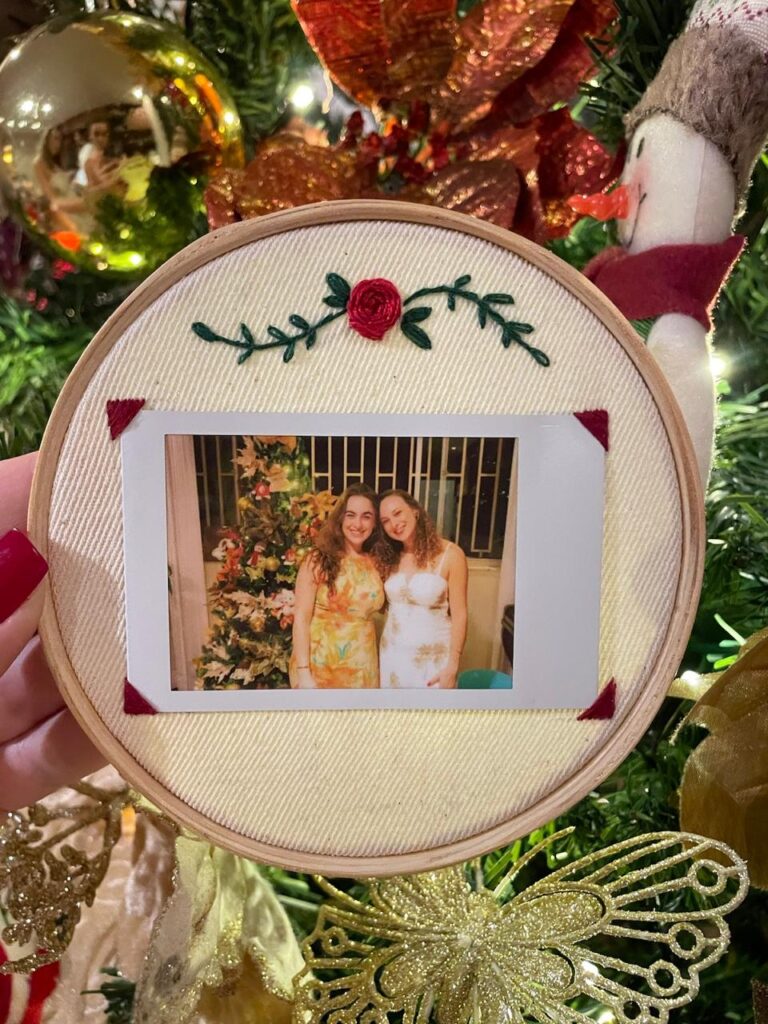 Bastidor bordado com flor vermelha e foto de duas jovens sorrindo, representando a história afetiva por trás da marca de moda infantil Mares que Bordam.